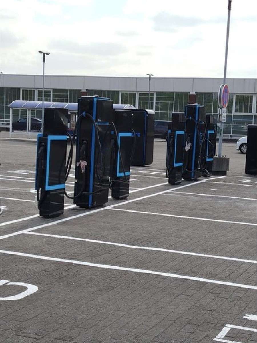 Bornes de recharge rapide en courant continu alignées sur les places d'un parking extérieur, câbles de charge en place, habillage encore protégé par son film de pose.